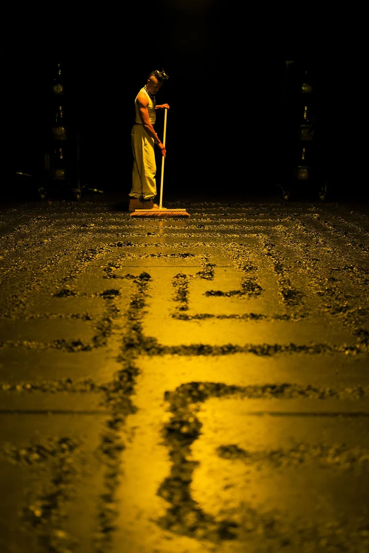 A performer holds a broom at the end of a labyrinth traced across a yellow-lit floor.