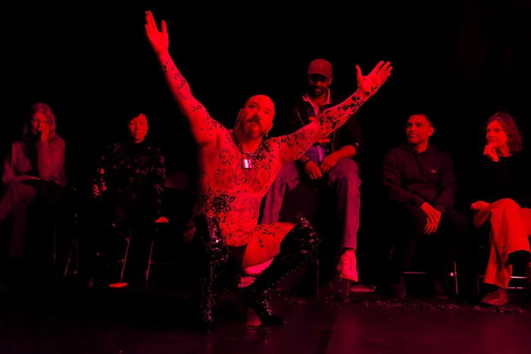 A performer kneels with raised arms in red light, surrounded by seated audience members.